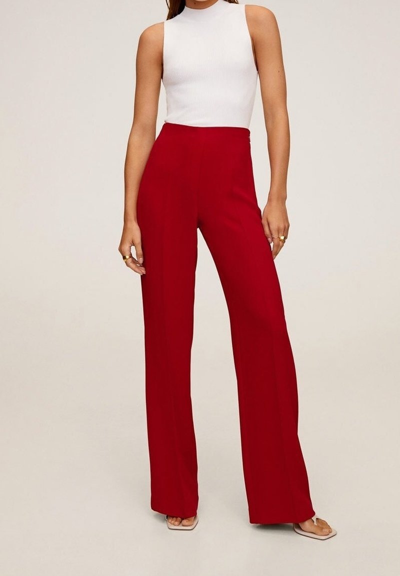 Femme portant un pantalon rouge taille haute à jambes larges et un top sans manches blanc à col montant, debout sur un fond beige clair.