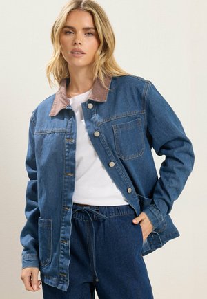 Młoda kobieta ubrana w niebieską dżinsową kurtkę z brązowym kołnierzem na białej koszuli i dopasowane dżinsy, stoi z jedną ręką w kieszeni.