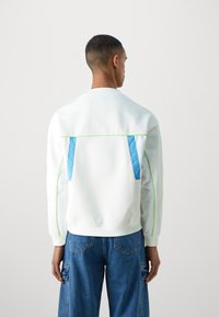 Sweat à capuche blanc avec des accents bleus et verts, présentant une coupe décontractée, un col rond et des poches à l'arrière. Associe à un jean cargo bleu large.