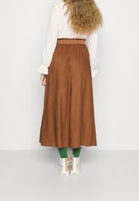 Donna con lunghi capelli ricci che indossa una blouse bianca, una gonna midi marrone, calzini verdi e sneakers Converse bianche, di spalle.