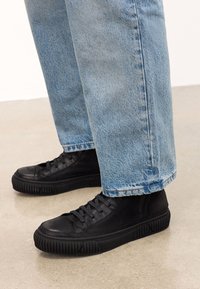 Mustad musta nahast kõrged tossud, millel on tekstureeritud pind ja paks kummitald, kanda helesiniste, lõdvalt istuvate teksadega.