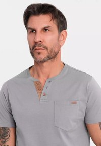 Camisa de manga corta gris con una tapeta abotonada marrón y bolsillo. Presenta una textura suave y un diseño simple y limpio.