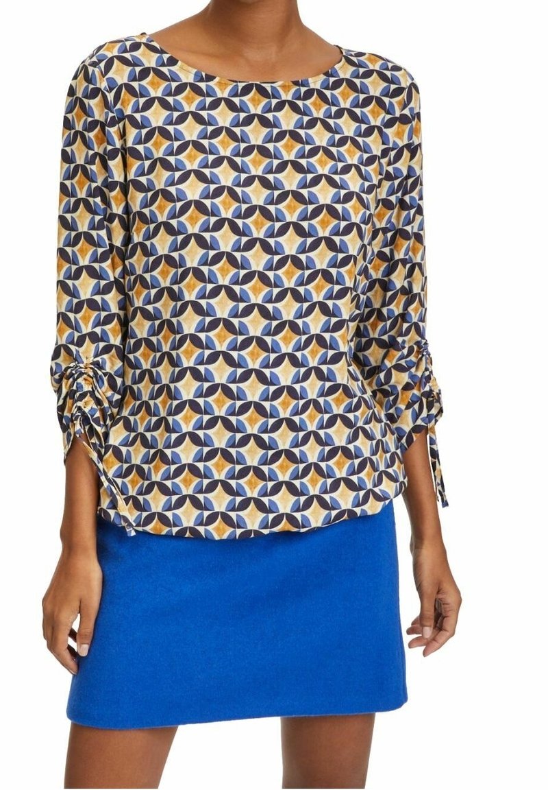 Blusa stampata multicolore con maniche a tre quarti, caratterizzata da un colletto rotondo e polsini arricciati. Abbinata a una mini gonna di feltro blu.