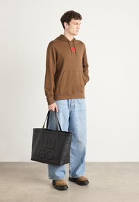 Brun hette med en frontlomme og rød logo-patch, kombinert med lyseblå vide jeans og en svart lær tote-bag med preget skrift.