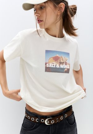 Crème katoenen t-shirt met een rechthoekige afbeelding met "SALT & WIND" over een afbeelding van een schelp, gecombineerd met zwarte high-waisted jeans.