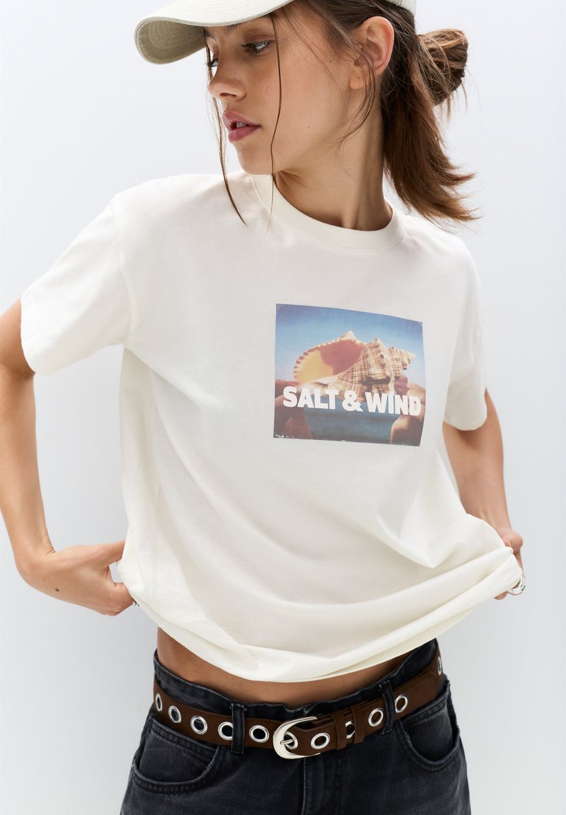 Cremefarvet bomulds t-shirt med et rektangulært grafisk tryk med "SALT & WIND" over et billede af en musling, parret med sorte højtaljede jeans.