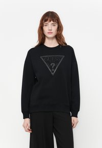 Mujer con cabello rojo de longitud media vistiendo un sudadera negra de Guess con un gran logo triangular y pantalones negros, de pie contra un fondo blanco.