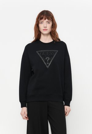Donna con capelli rossi di media lunghezza che indossa un felpa nera di Guess con un grande logo triangolare e pantaloni neri, in piedi contro uno sfondo bianco.