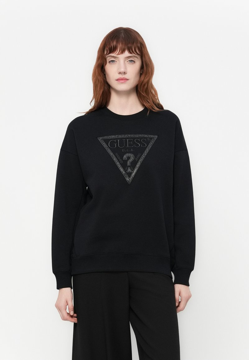 Mujer con cabello rojo de longitud media vistiendo un sudadera negra de Guess con un gran logo triangular y pantalones negros, de pie contra un fondo blanco.