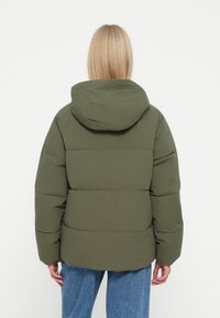 Chaqueta acolchada verde con capucha, de tela texturizada, forma cuadrada y detalles de acolchado segmentado. Combinada con jeans de mezclilla azul claro.