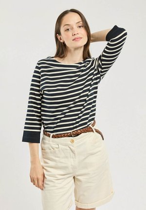 Jeune femme portant un haut à manches longues rayé noir et blanc, un short beige roulé et une ceinture tressée marron, posant avec une main derrière la tête.