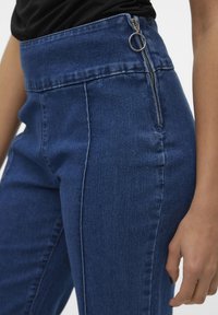 Visoke jeans hlače iz temno modrega jeansa, ki imajo zadrgo na strani in obročni poteg, z gladko teksturo in krojem, ki se prilega nogi.