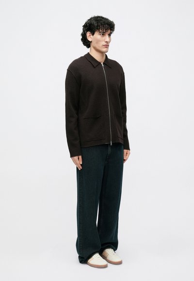 Brązowy sweter na zamek z kołnierzykiem, dwiema przednimi kieszeniami, w zestawieniu z ciemnymi dżinsami o szerokich nogawkach i białymi trampkami. Gładka faktura na całej powierzchni.