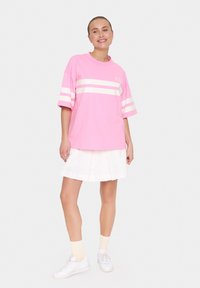 T-shirt oversize rosa con strisce orizzontali bianche e logo, abbinata a una gonna plissettata bianca e sneakers bianche, con calzini leggeri.