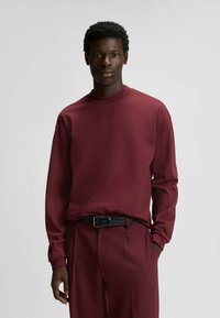 Bordeaux langemouwen shirt met een ronde hals en subtiele logodetail; gecombineerd met bijpassende broek en een zwarte riem. Gladde stofstructuur.