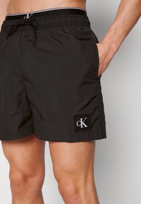 Shorts de bain noirs en matériau léger, avec une taille élastique à cordon de serrage et un petit patch logo cK sur le côté.