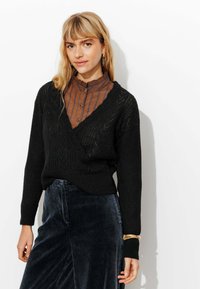 Un pull châle en tricot noir avec un motif texturé, superposé sur une chemise à boutons rayée marron. Le mannequin porte un pantalon en velours côtelé bleu marine.