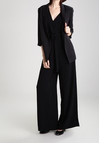 Svart byxdress med vida ben, djup V-ringning, bälte i midjan och en matchande lätt blazer. Slät textur, minimalistisk design.