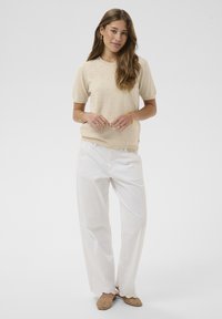 Naine, kes kannab beežikat lühikeste varrukatega kootud topi, valgeid laiade säärtega pükse silmatorkavate äärtega ja beežikaid kootud baleriinikingi, seisab valge tausta ees.