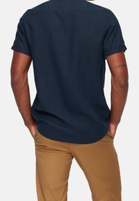 Camicia a maniche corte di colore blu navy scuro realizzata in tessuto testurizzato, con colletto a bottoni e orlo curvo. Abbinata a pantaloni beige aderenti.