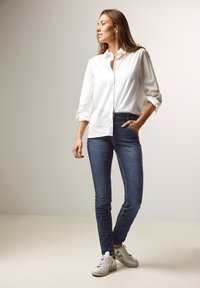 Chemise blanche à manches longues avec des manches roulées, associée à un jean skinny bleu foncé et des baskets grises, se tenant sur un fond clair.