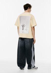 Gul oversized t-shirt med et sort-hvidt håndgrafik på ryggen. Bæres sammen med brede denim bukser i nuancer af mørk og lys grå.