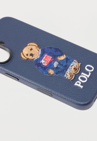 Θήκη τηλεφώνου από δέρμα σε ναυτικό μπλε χρώμα με κεντητό αρκουδάκι που φοράει πουλόβερ και τζιν, με το κείμενο "POLO" στο κάτω μέρος. Λεπτές υφές στη δομή.