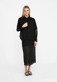 Zwarte, doorschijnende blouse met knopen en lange mouwen, gedragen over een zwarte maxi-rok met borduursels. De schoenen zijn zwarte platte schoenen met een textuureffect.