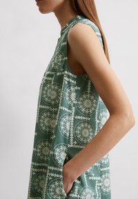 Grön ärmlös klänning med geometriskt vitt mönster, hög halsringning och fickor på sidorna. Mjukt tyg.