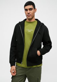 Μαύρη φούτερ ζακέτα με φερμουάρ και εμπρόσθιες τσέπες, σε συνδυασμό με πράσινο t-shirt από Calvin Klein. Το μοντέλο φοράει ελιά πράσινο παντελόνι.