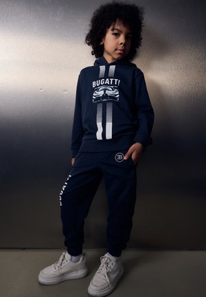 Bambino con capelli ricci che indossa una felpa con cappuccio Bugatti blu navy e pantaloni della tuta coordinati, in posa davanti a un muro metallico con scarpe da ginnastica bianche.