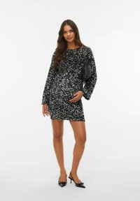 Zwarte paillettenjurk met lange mouwen, getailleerde pasvorm en ronde halslijn. Gepaard met zwarte puntige hakken en een zwarte tas.