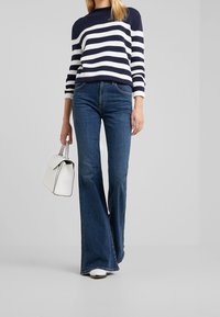 Blaue Schlagjeans mit einer dunklen Waschung, kombiniert mit einem navy- und weiß gestreiften Strickpullover und einer weißen Handtasche mit strukturiertem Design.