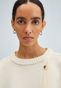 Cream knit sweater with a ribbed neckline, featuring a side button accent. Smooth texture with a loose fit and minimal design details.
