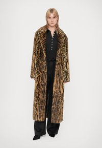Casaco de pêlo sintético com padrão de leopardo, de design longo, com um colarinho largo e frente de dupla abotoadura. Usado sobre uma blusa preta e calças.