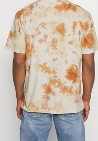 Zadní pohled na osobu v tričku s krátkým rukávem ve stylu tie-dye s oranžovými a béžovými vzory, doplněné světle modrými džíny.