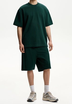 Homme portant un t-shirt oversize vert foncé et un short long assorti, avec des chaussettes blanches et des baskets beige, debout devant un fond uni.
