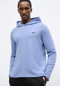 Sweatshirt azul claro com capuz, tecido texturizado, mangas compridas e logótipo preto bordado no peito. Combinado com calças pretas.