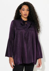 Blusa viola realizzata in tessuto lucido, con un fiocco al collo, maniche a campana lunghe e un taglio morbido.