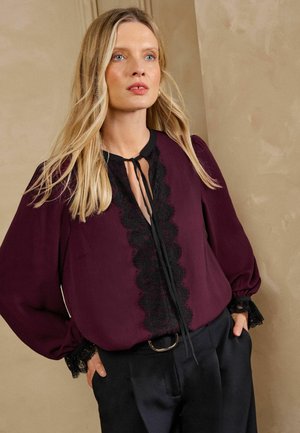 Vrouw met lang blond haar, die een paarse blouse draagt met zwarte kanten rand en zwarte broek, staat met handen in de zakken.