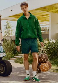 Veste zippée verte avec texture côtelée, shorts assortis, chaussettes blanches, et baskets vertes. Tient un sac en filet de ballons de basket.