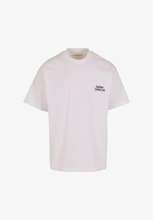 T-shirt en coton blanc à col rond et manches courtes. Présente un petit texte noir sur le côté supérieur gauche : « Another Cotton Lab. »