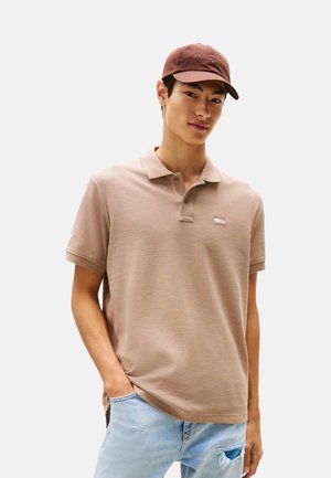 Polo beige con acabado texturizado, con una tapeta de tres botones y un pequeño logotipo. Combinado con una gorra marrón. Jeans de mezclilla clara.