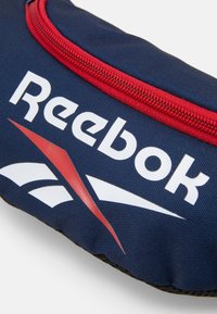 Reebok torbica koja ima tamnoplavu tkaninu s crvenim patentnim zatvaračem, bijelim logotipom i crvenom trakastom dekoracijom blizu baze.