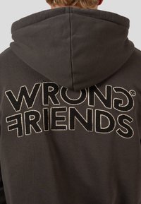 Tmavě šedá mikina s kapucí, na které je vzadu velký kontrastní nápis "WRONG FRIENDS" vyšitý v černé a bílé barvě. Disponuje prostornou kapucí.
