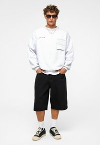 Sweatshirt gris clair avec une poche poitrine, associé à un short en denim noir. Le modèle porte des lunettes de soleil noires et des baskets noires avec des accents blancs.