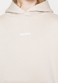 Sweat à capuche beige clair avec un petit logo blanc "WRSTBHVR" brodé sur la poitrine, porté par une personne aux cheveux blonds.