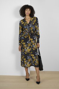 Femeie cu păr creț, îmbrăcată într-o rochie florală midi neagră cu flori galbene și gri, ținând o geantă de mână neagră, stând pe un podea din lemn.