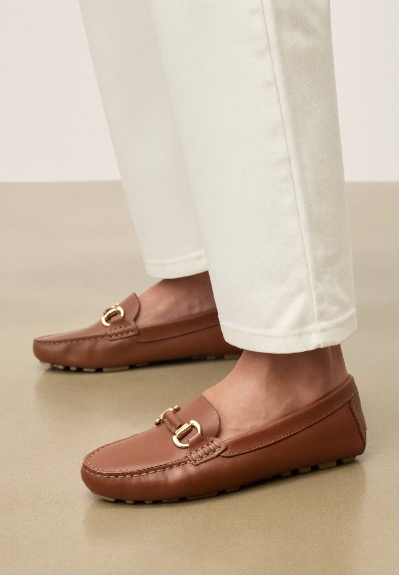 Mocassins en cuir marron avec détail de boucle dorée portés avec un pantalon blanc sur un sol beige.