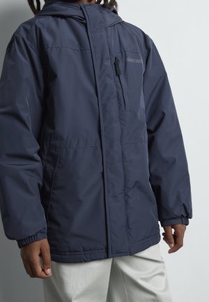 Mand iført marineblå hættetrøje med lynlås på brystlomme og elastiske manchetter, kombineret med lyse bukser.